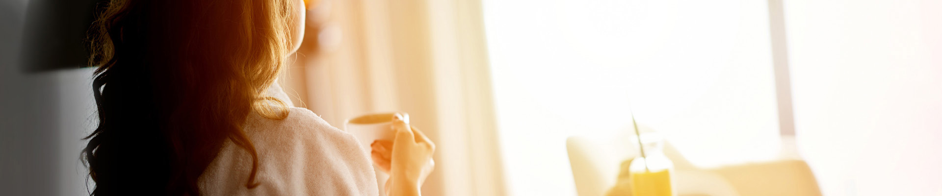 Woman looking out sunny hotel window drinking coffee